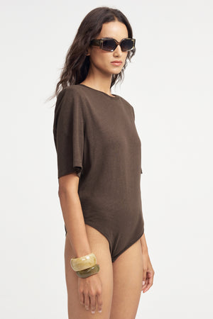 Woman wearing a premium brown linen tee bodysuit with sunglasses on a grey background