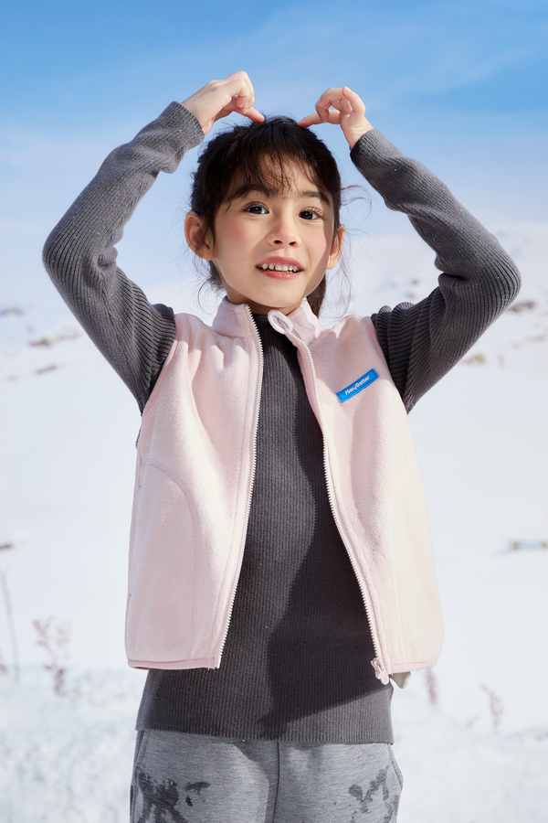 A girl in a gray sweater and pink soft lightweight vest standing in a snowy landscape with a blue sky.